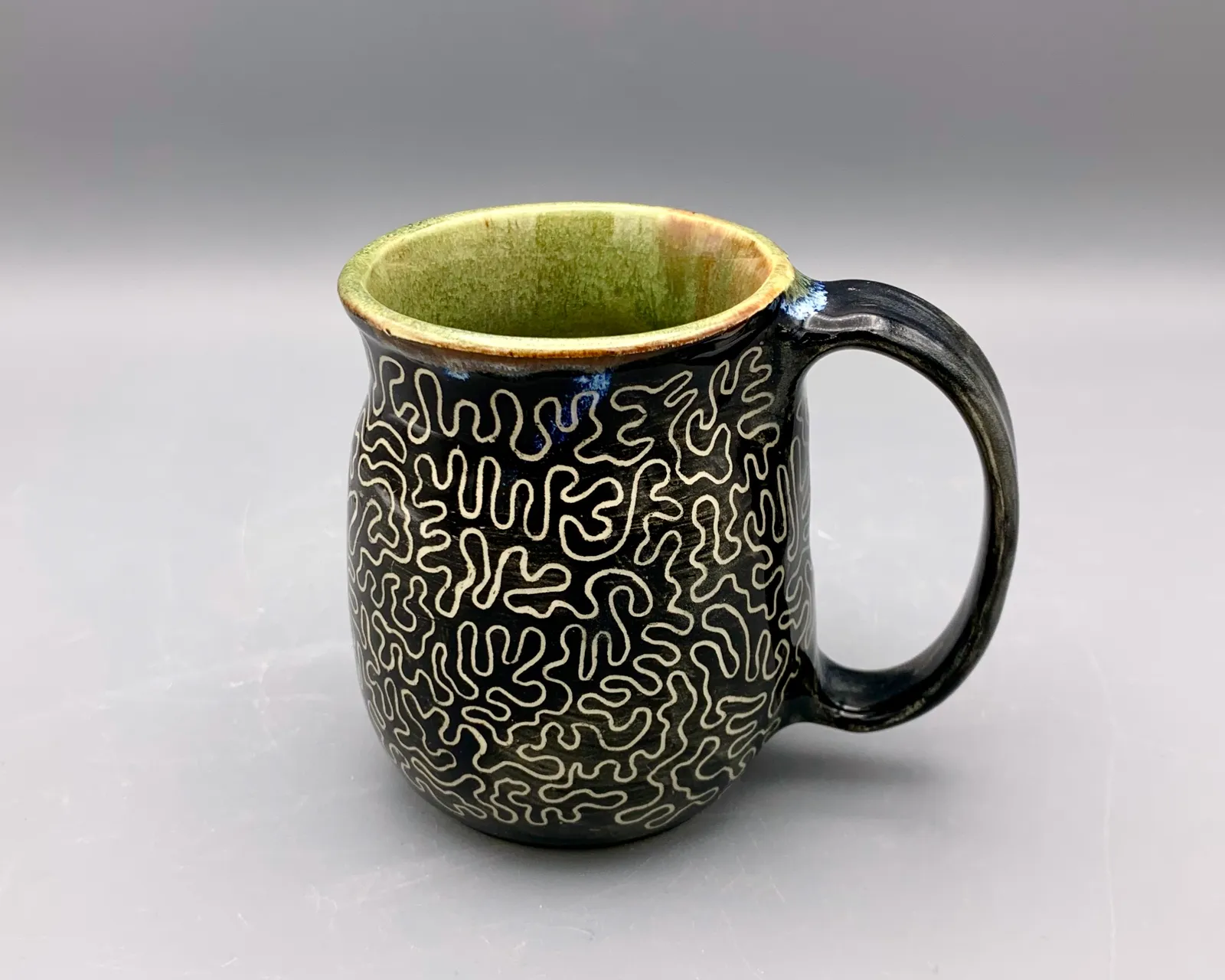 Sgraffito mug, 2024, glazed stoneware fired to Cone 6 oxidation, approx. 4 x 5 x 4 in. A black mug with white carving in a squiggly pattern.