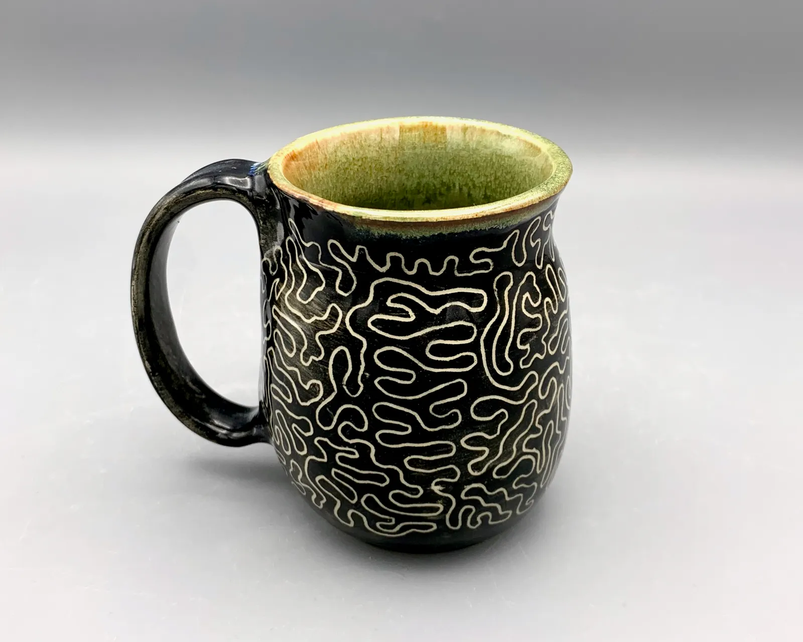 Sgraffito mug, 2024, glazed stoneware fired to Cone 6 oxidation, approx. 4 x 5 x 4 in. A black mug with white carving in a squiggly pattern.