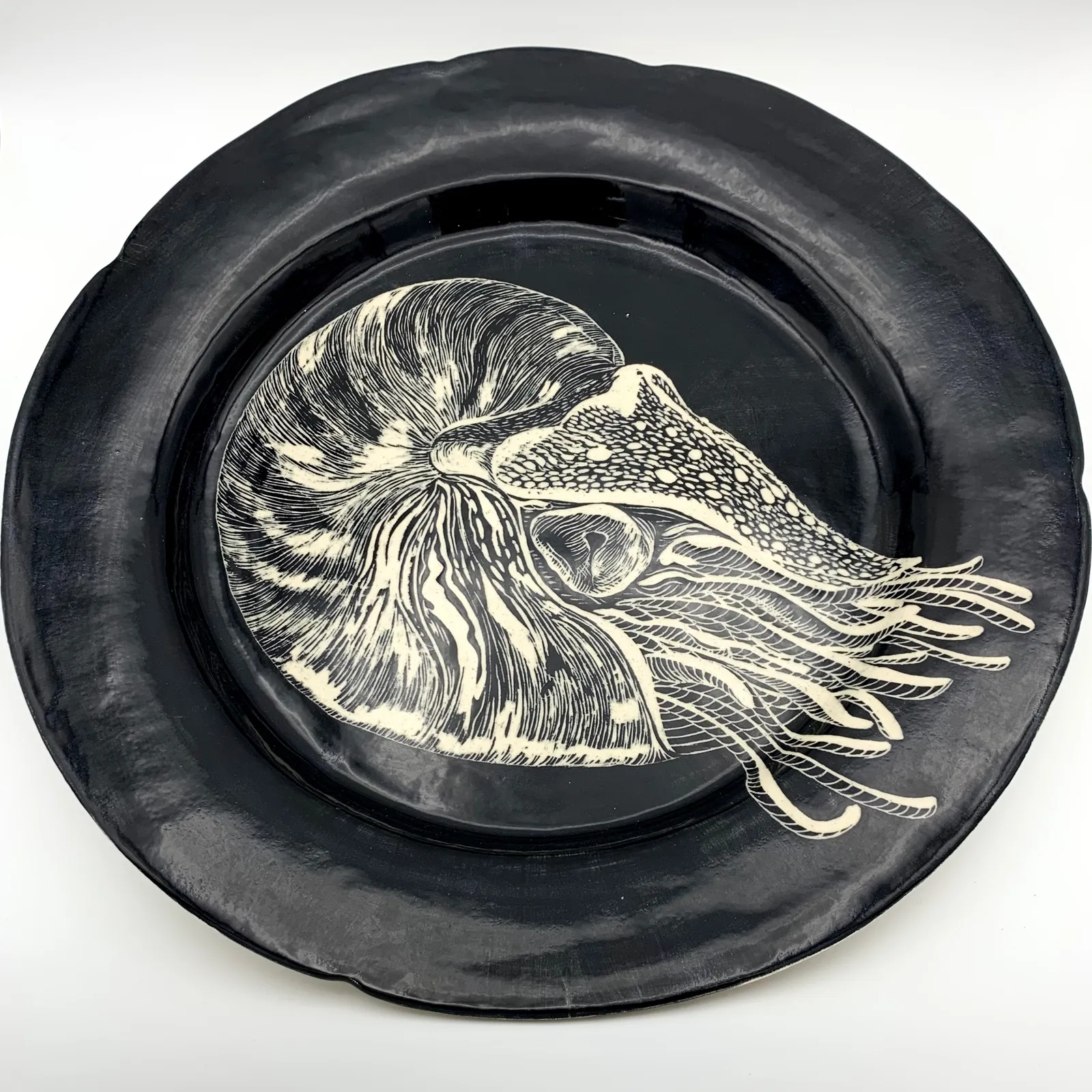 Sgraffito platter, 2021, glazed stoneware fired to Cone 6 oxidation, approx. 1 x 14 x 14 in. A black platter with white carving of a nautilus.