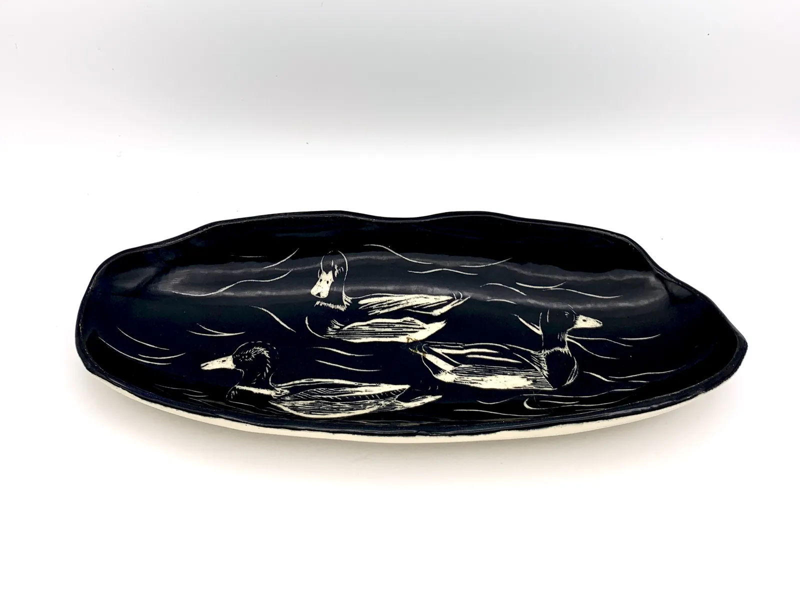 Sgraffito dish, 2019, glazed stoneware fired to Cone 6 oxidation, approx. 1 x 9 x 5 in. A black dish with white carving of ducks on water.