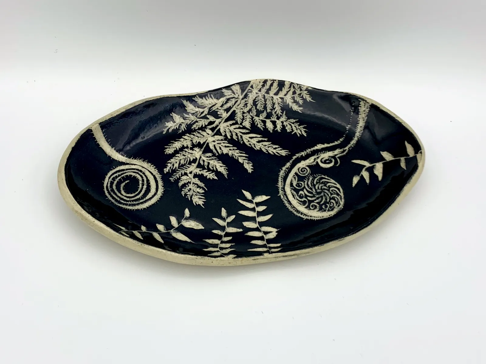 Sgraffito dish, 2019, glazed stoneware fired to Cone 6 oxidation, approx. 1 x 9 x 6 in. A black oval dish with white carving in a fern pattern.