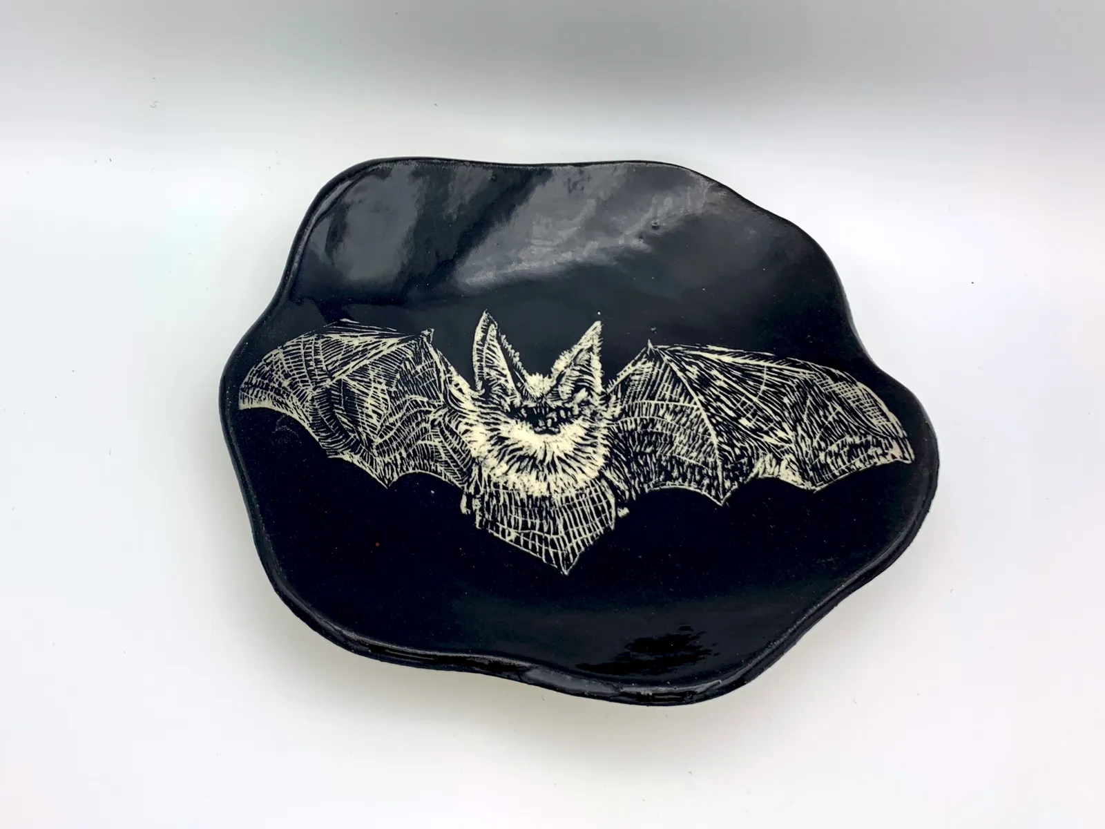 Sgraffito bat dish, 2019, glazed stoneware fired to Cone 6 oxidation, 2 x 8 x 7 in. A black dish with white carving of a bat.