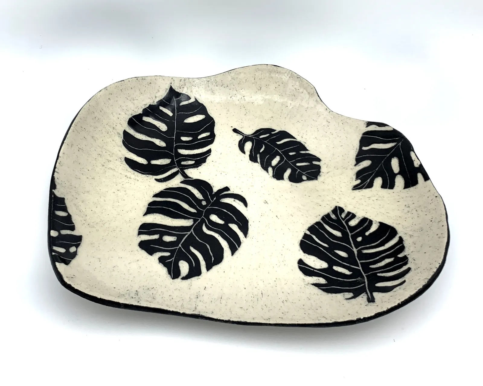 Sgraffito dish, 2019, glazed stoneware fired to Cone 6 oxidation, approx. 1 x 10 x 7 in. A black freeform dish with white carving in a monstera leaf pattern.