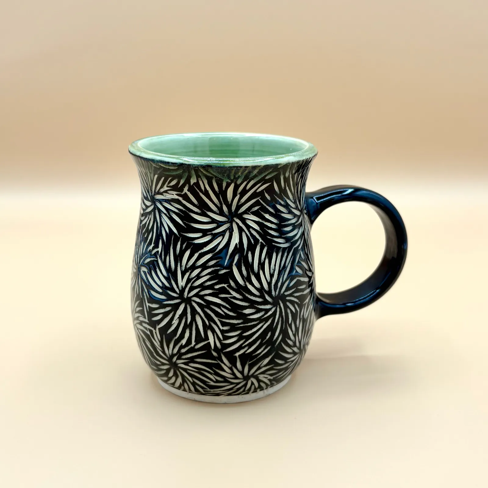 Sgraffito mug, 2026, glazed stoneware fired to Cone 6 oxidation, approx. 4 x 5 x 4 in. A black mug with white carving in a floral pattern.