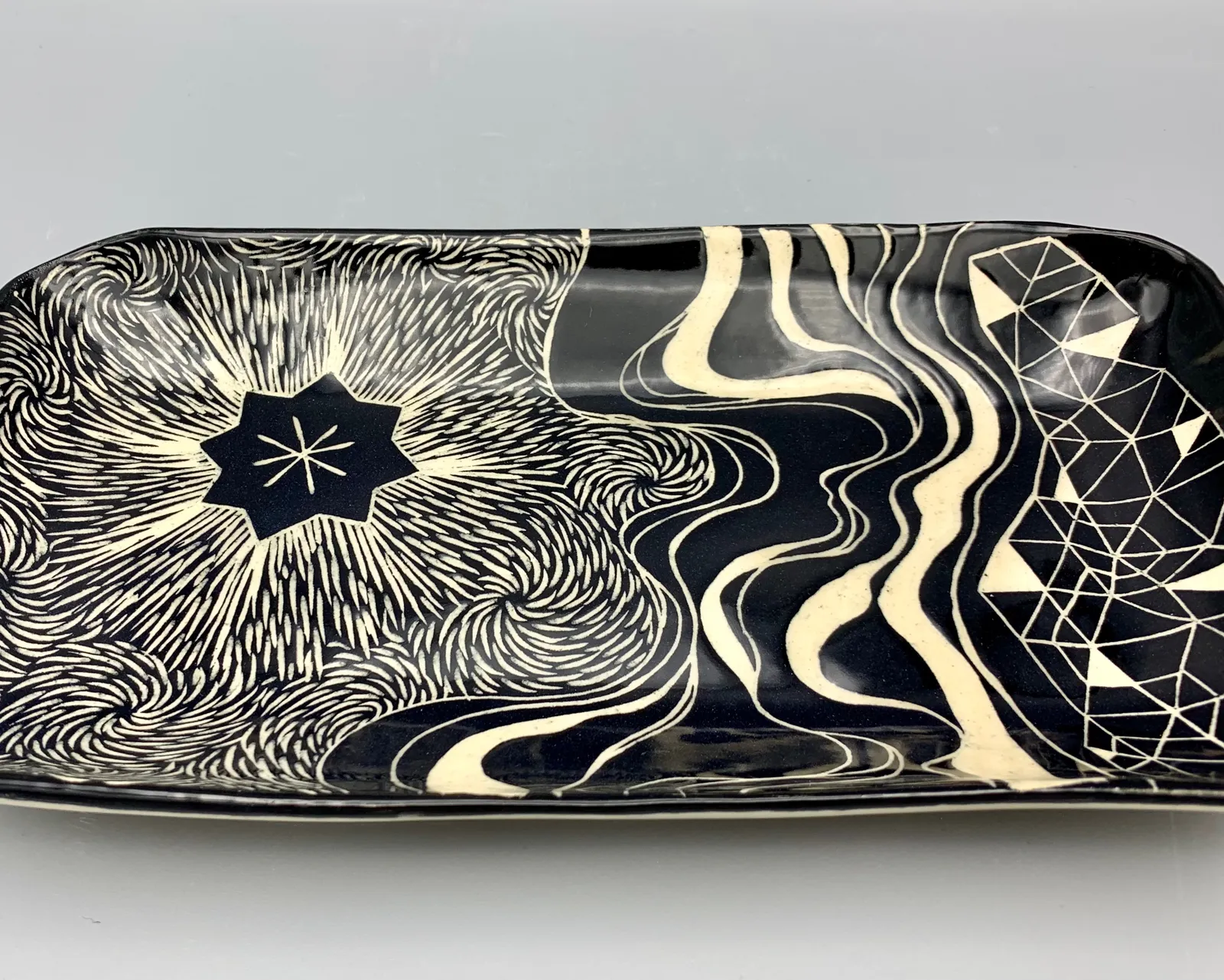 Sgraffito dish, 2021, glazed stoneware fired to Cone 6 oxidation, approx. 1 x 12 x 6 in. A black rectangular dish with white carving in a variety of patterns.