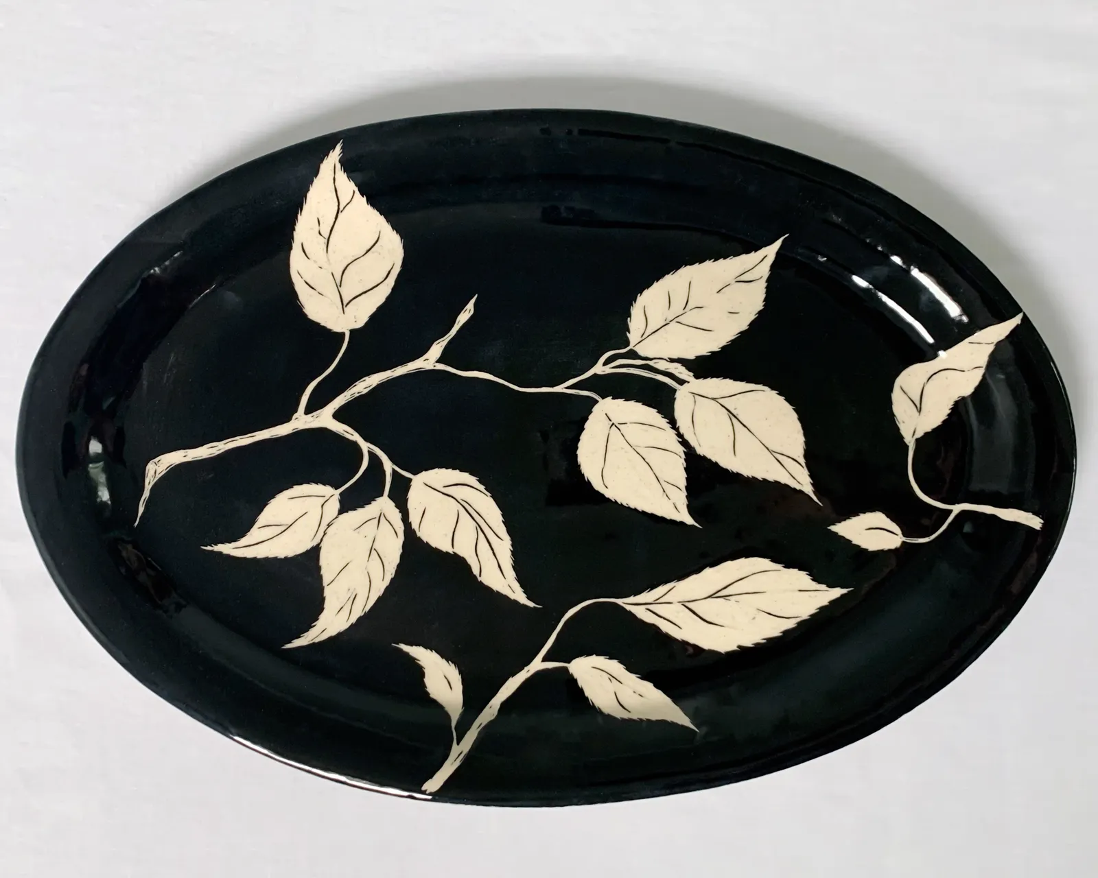Sgraffito platter, 2021, glazed stoneware fired to Cone 6 oxidation, approx. 1 x 16 x 10 in. A black platter with white carving in a leaf pattern.